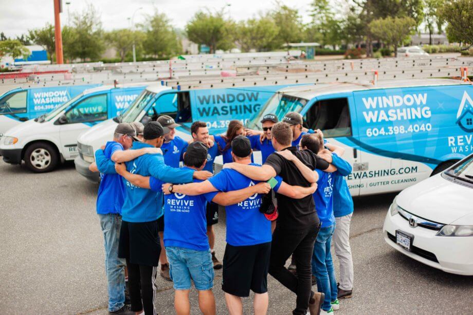 Team of window cleaning employees standing in front of work vans with their arms linked around each others shoulders in a circle.